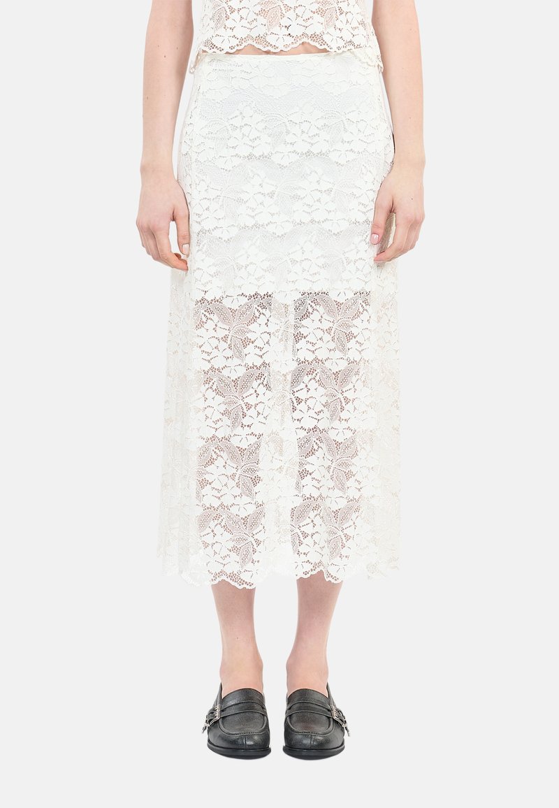 Jupe midi en dentelle blanche avec un motif floral, texture transparente et ourlet festonné. Des mocassins noirs avec des éléments en métal argenté complètent la tenue.