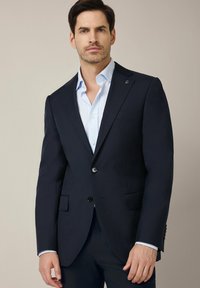 Granatowa marynarka dopasowana, z dwoma guzikami, klapami notcha oraz bocznymi kieszeniami. Pod spodem widoczna jasnoniebieska koszula o gładkiej fakturze materiału.