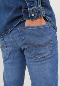 Ruka zasunutá do zadní kapsy modrých džínů z denimu, viditelný je také rukáv modré džínové košile, která ladí s kalhotami.