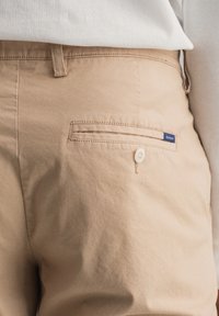 Pantalon beige clair en mélange de coton, avec une poche arrière munie d'un bouton et un accent cousu. Petite étiquette avec le nom de la marque visible.