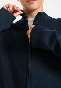 Mörkblå zip-up-tröja med ribbade muddar, hög krage och dragkedja. Mjuk textur, lätt material.