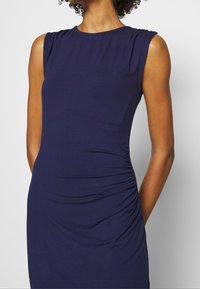 Femme portant une robe bleu marine sans manches avec un détail froncé sur le côté, debout devant un fond clair uni.