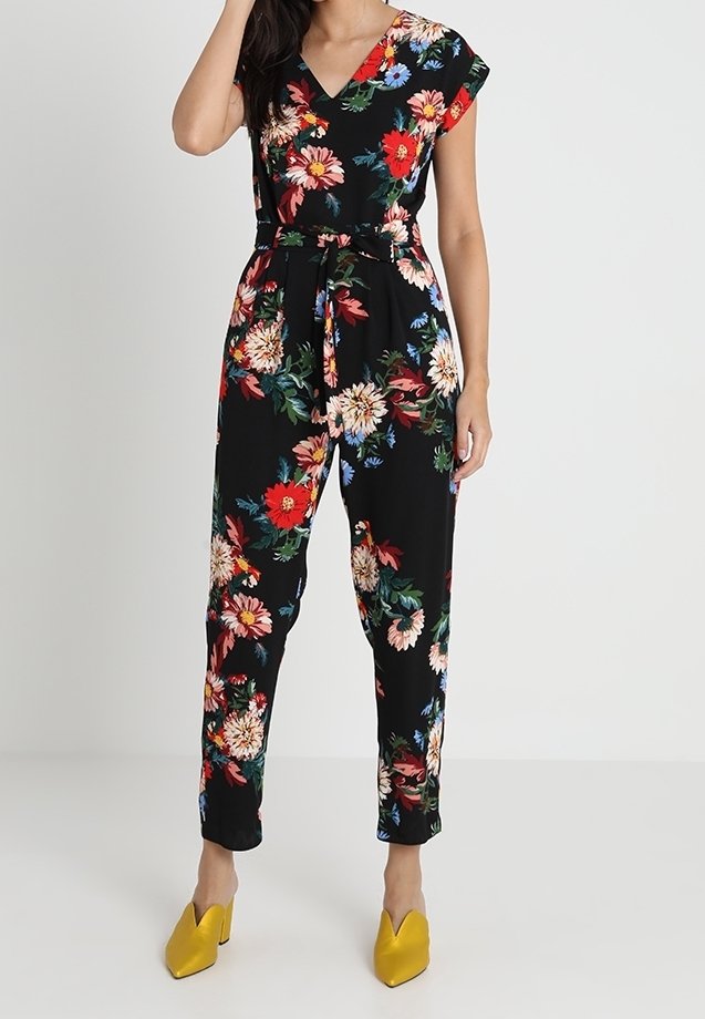 Combinaison florale avec un fond noir, manches courtes, encolure en V, taille nouée, ornée de fleurs rouges, jaunes et bleues. Portée avec des talons jaunes.