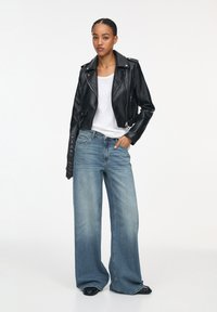 Zwarte leren motorjack met zilveren ritsen, gecombineerd met wijdvallende lichtblauwe jeans. Model draagt een wit T-shirt eronder.
