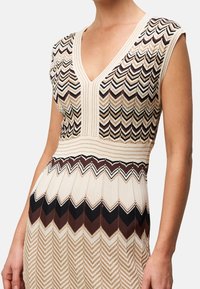 Sukienka dziana z dekoltem w serek, ozdobiona zygzakowatym wzorem w kolorach kremowym, brązowym i czarnym. Krótkie rękawy i teksturowane detale na całej powierzchni.