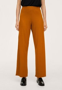 Pantalon à jambes larges en tissu texturé couleur orange brûlé. Il présente une taille haute et un ourlet effiloché. Porté avec des mocassins noirs.