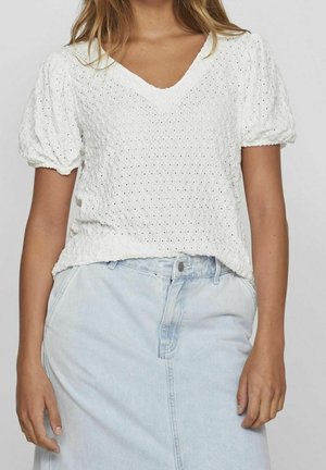 Kobieta ubrana w biały, teksturowany top z krótkim rękawem i dekoltem w serek, zestawiony z jasnoniebieską, wysokim stanem spódnicą dżinsową.