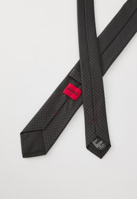 Cravate en soie texturée gris foncé avec une étiquette rouge Hugo et une étiquette d'entretien, posée à plat sur une surface blanche.