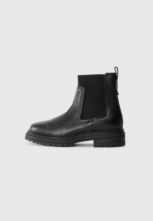 Bottine en cuir noir avec panneau latéral élastique côtelé, languette à l'arrière et semelle épaisse texturée, montrée de profil sur fond uni.