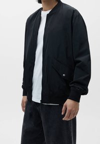 Chaqueta bomber negra con cierre de cremallera en el frente, puños acanalados y bolsillos laterales. Llevada sobre una camiseta blanca, combinada con jeans oscuros y holgados.