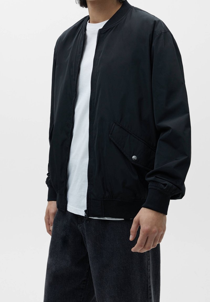 Chaqueta bomber negra con cierre de cremallera en el frente, puños acanalados y bolsillos laterales. Llevada sobre una camiseta blanca, combinada con jeans oscuros y holgados.