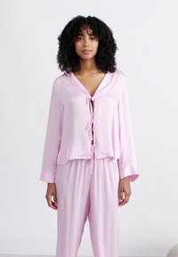 Γυναίκα με σγουρά μαλλιά, φορώντας ροζ πυτζάμα με δετό μπροστινό μέρος και αντίστοιχο φαρδύ παντελόνι, στεκόμενη μπροστά σε απλό λευκό φόντο.