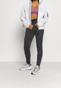 Mujer vestida con una chaqueta blanca con cremallera, sujetador deportivo mauve, pantalones jogger gris oscuro y zapatillas gris claro con acentos coral, de pie sobre un suelo liso.