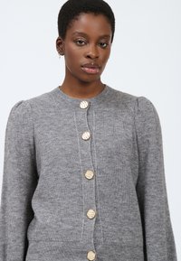 Cardigan grigio con scollatura tonda, maniche lunghe, tessuto testurizzato e sei bottoni dorati a strisce sulla parte anteriore.