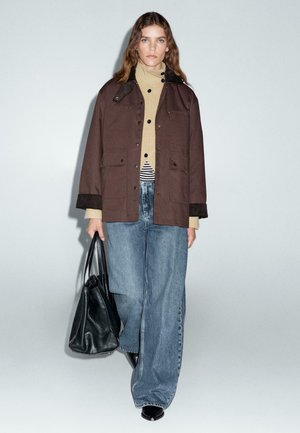 Veste marron avec un design boutonné, col roulé beige, chemise rayée, jean bleu en denim à jambes larges et un sac fourre-tout noir. Textures lisses.