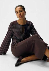 Jonge vrouw zittend op de vloer, gekleed in een paarse geruite blouse, een bruine rok en zwarte puntige flats, tegen een witte achtergrond.