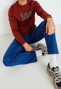 Červená mikina s bílým logem "Lee", modré džíny, šedé a černé sportovní boty, posezení na světlém pozadí.
