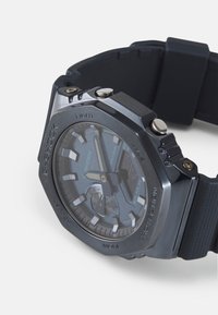Ceas G-SHOCK cu un carcasă octogonală metallică închisă, cadran albastru, accente argintii și o brățară din cauciuc negru. Dispune de afișaje numerice și analogice.