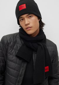 Black ribbed knit beanie and scarf with red "HUGO" logo; worn with a black puffer jacket featuring a zip closure and quilted texture.