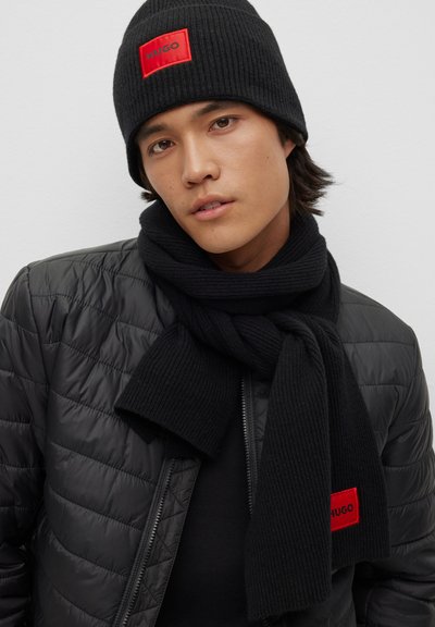 Bonnet en tricot côtelé noir et écharpe avec le logo rouge "HUGO" ; portée avec une doudoune noire à fermeture éclair et texture matelassée.
