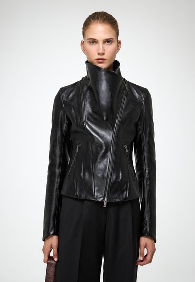 Femme avec les cheveux attachés portant une veste en cuir noire brillante avec un col zippé asymétrique haut et un pantalon noir à jambes larges.
