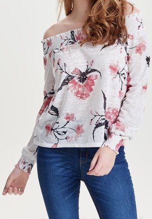 Femme portant un haut blanc épaules dénudées avec un imprimé floral rose et noir et un jean bleu, avec des cheveux bruns bouclés sur une épaule.