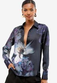 Femeie îmbrăcată cu o bluză din satin negru, cu imprimeuri mari de flori purpurii și albe, cu manșete cu nasturi, asortată cu pantaloni negri.