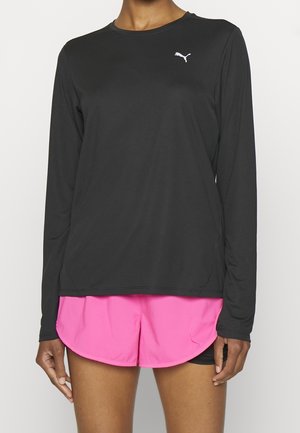 Osoba má na sobě černý dlouhý rukáv Puma a jasně růžové běžecké šortky, stojí před jednobarevným pozadím.