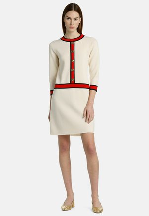 Strickkleid in Creme mit dreiviertel Ärmeln, roten und schwarzen Akzenten, goldenen Knöpfen entlang der Vorderseite und einem schwingenden Saum. Goldene Ballerinas runden den Look ab.