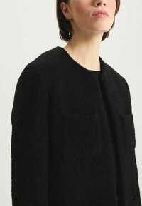Cardigan en laine noir texturé avec un col rond, deux poches avant et fermeture par boutons, présenté de la poitrine supérieure vers le haut.