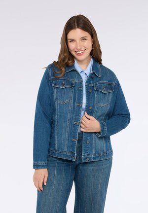Veste en denim de couleur bleu foncé, avec une fermeture à boutons sur le devant, deux poches poitrine et des détails de coutures verticales. Accompagnée d'une chemise bleu clair.