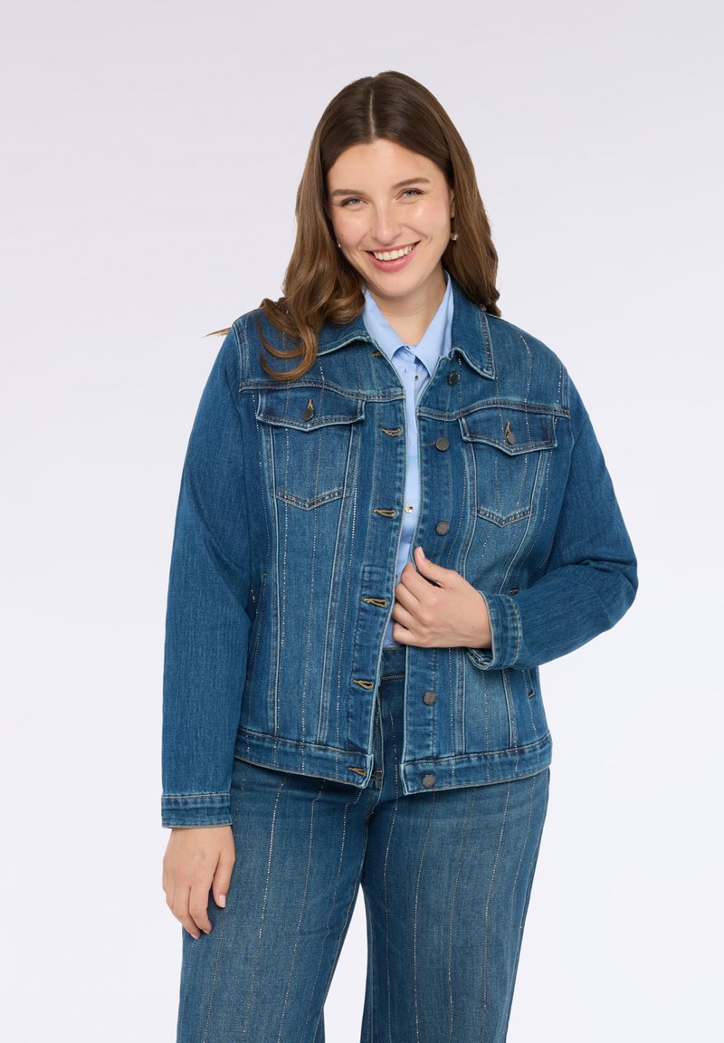 Jachetă din denim cu o culoare albastru închis, cu închidere cu nasturi, două buzunare pe piept și detalii de cusătură verticale. Însoțită de o cămașă albastră deschisă.