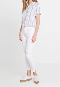 Camicia a maniche corte con bottoni a righe blu, abbinata a jeans skinny bianchi strappati e sneakers bianche con suola in juta.