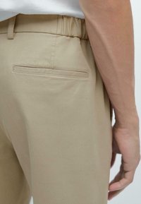 Pantalons beiges en tissu lisse, avec une taille élastique et une poche arrière simple avec un détail de couture plate.