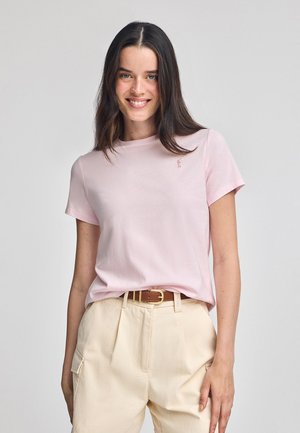 Camiseta de algodón rosa claro con mangas cortas, cuello redondo y un pequeño logo bordado en el lado izquierdo del pecho. Combinada con pantalones beige.