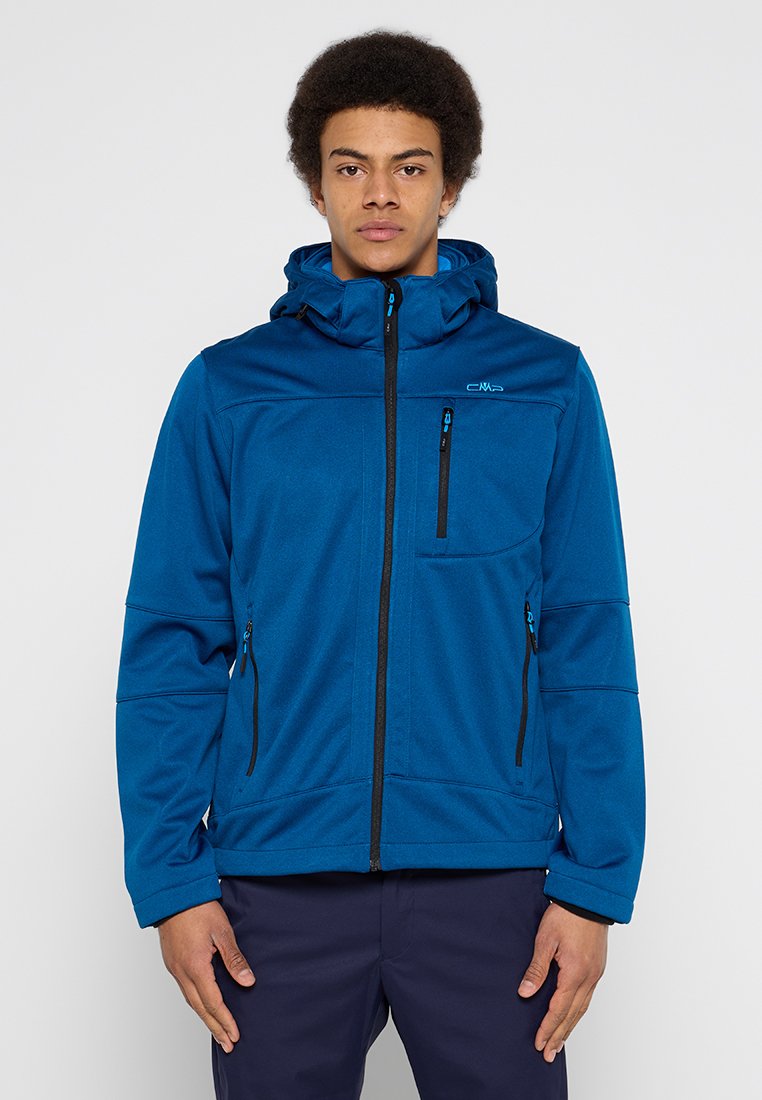 Joven vestido con una chaqueta con capucha azul y cremallera, con bolsillos en el pecho y laterales con cremallera, de pie contra un fondo claro y liso.