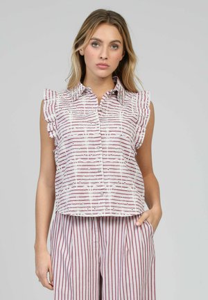 Femme portant un chemisier sans manches à col avec des épaules à volants et un pantalon rayé assorti, debout devant un fond uni.