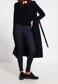 Marinblå lång kappa över en svart tröja, mörka jeans och svarta skor. Har en krage, smal passform och en jämn textur genom hela plagget.