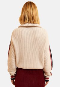 Pull en maille côtelée beige avec un col montant et des accents rayés multicolores sur les manches ; associé à une jupe en velours côtelé rouge foncé.