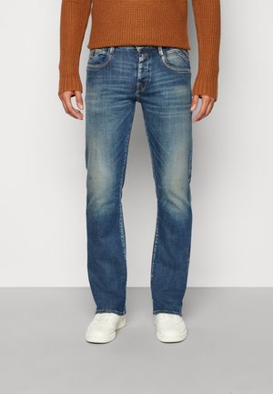 Uomo con jeans blu slavati, scarpe da ginnastica bianche e maglione color ruggine, in piedi davanti a uno sfondo semplice grigio e bianco.