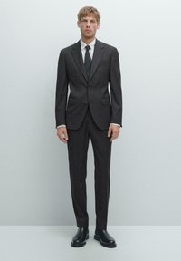 Zweiteiliger, dunkelgrauer Anzug mit Nadelstreifen und klassischem Revers, kombiniert mit einem weißen Hemd und einer dunklen Krawatte. Trägt schwarze Lederstiefel.