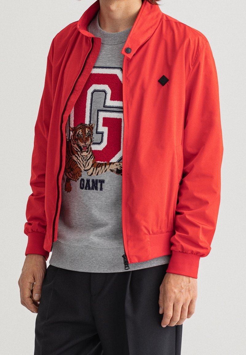 Veste bomber rouge avec col montant et poignets élastiques, portée sur un sweat-shirt gris clair orné d'une broderie de tigre et de lettres.