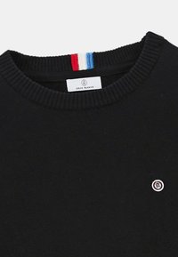 Pull noir à col rond avec col côtelé, petite patch logo rond sur la poitrine, et détail de rayures rouges, blanches et bleues à l'intérieur de l'encolure.