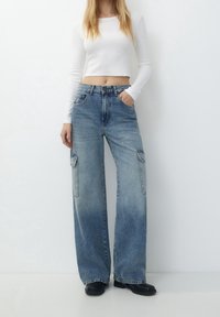 Breda ben blå denim cargo byxor med två framfickor, hög midja och subtila blekningar. Tillsammans med en åtsittande vit långärmad topp.