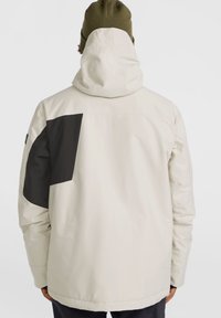 Veste imperméable beige clair avec un accent noir sur l'épaule gauche, dotée d'une capuche et de manches longues. Texture lisse et coupe décontractée.