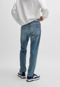 Vaaleansiniset farkut suoran leikkauksen omaavina, brändillä varustetulla vyötäröllä, kahdella takataskulla ja hienovaraisella kulumisella. Yhdistetty tummansinisiin lenkkareihin.