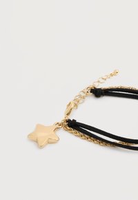 Χρυσό μενταγιόν σε σχήμα αστέρι, προσαρτημένο σε βραχιόλι με μαύρα κορδόνια και χρυσή αλυσίδα, σε ανοιχτό φόντο.