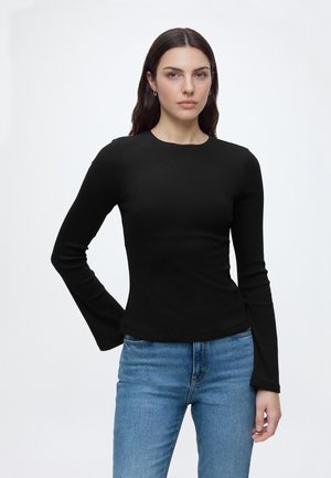 Femme aux longs cheveux foncés portant un haut noir ajusté à manches longues et un jean bleu, debout devant un fond clair uni.