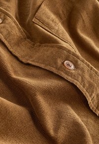 Tissu en velours côtelé marron avec des côtes visibles, un rabat de poche et un bouton marron cousu sur le vêtement.