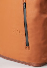 Borsa in pelle arancione con una cerniera nera sul lato e lettering in rilievo. Texture liscia con una base marrone a contrasto.